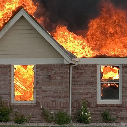 Brick house engulfed in flames with fire spreading across the roof and windows, producing thick black smoke. Home fire safety, fire prevention, and emergency response