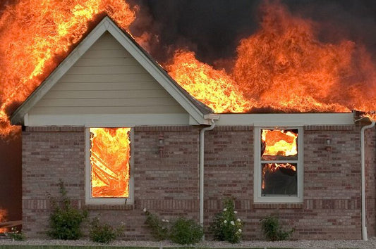 Brick house engulfed in flames with fire spreading across the roof and windows, producing thick black smoke. Home fire safety, fire prevention, and emergency response